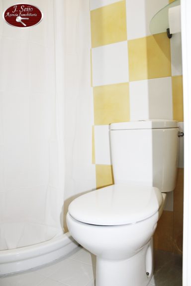 Baño con inodoro blanco y pared amarilla con azulejos. Cortina blanca en la ducha.